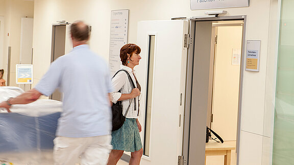 Eine Patientin geht in die Patientenaufnahme, ein Logistik-Mitarbeiter schiebt ein Patientenbett.
