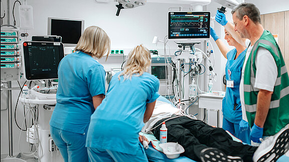 Vier medizinische Fachkräfte versorgen eine Person, die sich auf einer Krankenhausliege befindet, in einem modern ausgestatteten Behandlungsraum.