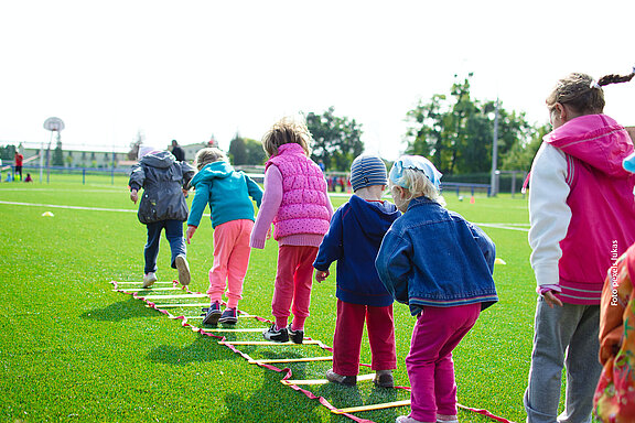 Kinder auf einer grünen Wiese in einer Reihe hintereinander machen ein Geschicklichkeits- und Hüpfspiel.
