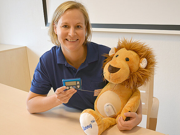 Oberärztin Nadine Scheffler hält ein Blutzuckermessgerät in der Hand, neben ihr am Tisch sitzt ein Kuscheltier (Löwe).