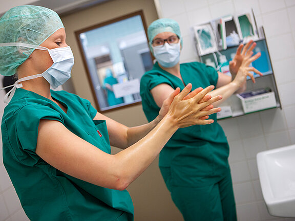 Zwei medizinische OP-Fachkräfte in grüner OP-Kleidung bereiten sich auf eine OP vor und desinfizieren Hände und Arme.