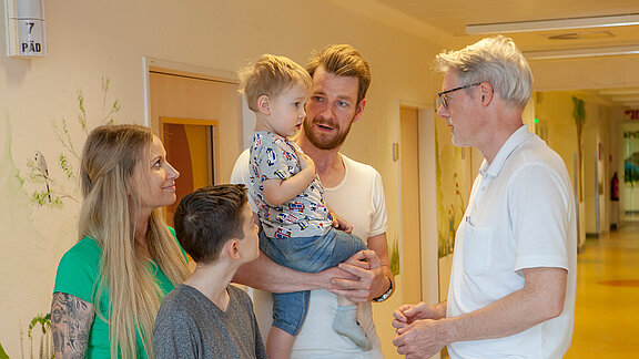 Chefarzt Dr. med. Georg Hillebrand (rechts) unterhält sich mit einem kleinen Patienten, der auf dem Arm des Vaters zusammen mit seiner Mutter und dem großen Bruder im Flur der Kinderklinik steht.