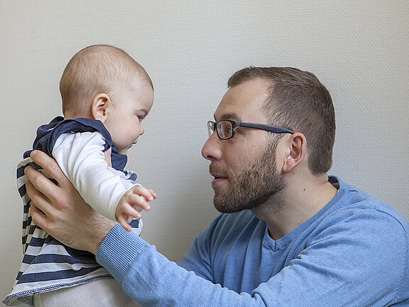 Ein junger Vater hält sein Baby hoch und blickt ihm in die Augen.