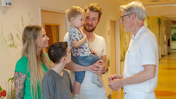 Chefarzt Dr. med. Georg Hillebrand (rechts) unterhält sich mit einem kleinen Patienten, der auf dem Arm des Vaters zusammen mit seiner Mutter und dem großen Bruder im Flur der Kinderklinik steht.