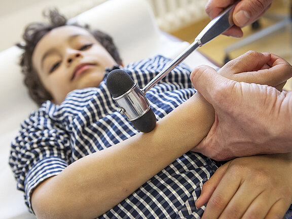 Ein Kind liegt auf einer Untersuchungsliege und wir von einem Arzt mit dem Reflexhammer untersucht.