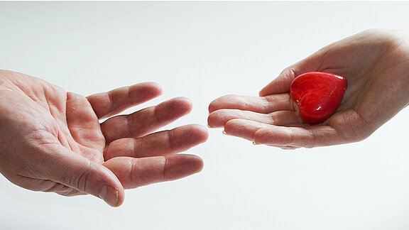 Zwei aufgehaltene Hände, auf der einen Hand liegt ein kleines rotes Herz.