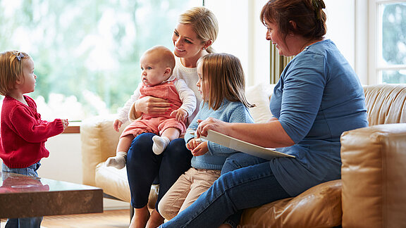Zwei Frauen sitzen mit drei Kindern unterschiedlichen Alters in einem hellen Wohnzimmer