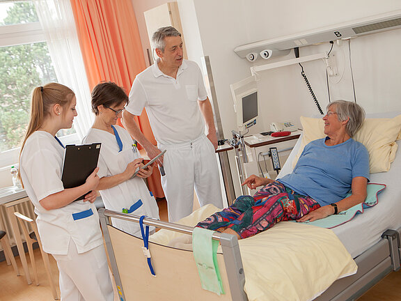 Chefarzt Prof. Dr. med. Michael Neipp und zwei medizinische Fachkräfte im Gespräch mit einer Patientin am PatientInnenbett.