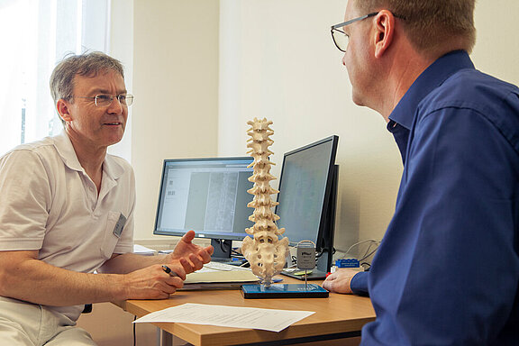 Dr. med. Wolfram Kluge klärt einen Patienten anhand eines Wirbelsäulenmodells über die SCS-Rückemankstimulation auf.