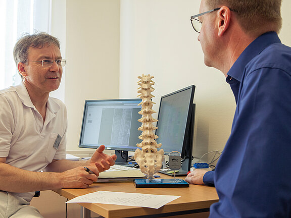 Dr. med. Wolfram Kluge klärt einen Patienten anhand eines Wirbelsäulenmodells über die SCS-Rückemankstimulation auf.