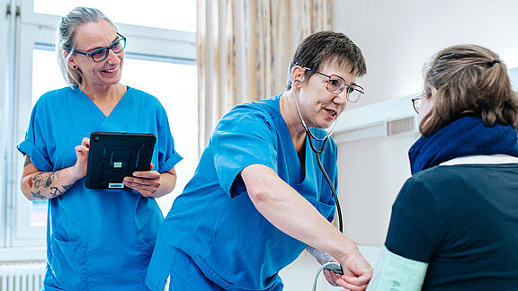 Zwei Pflegefachkräfte stehen am PatientInnenbett, die Patientin sitzt im Bett, eine Pflegefachkraft misst Blutdruck.