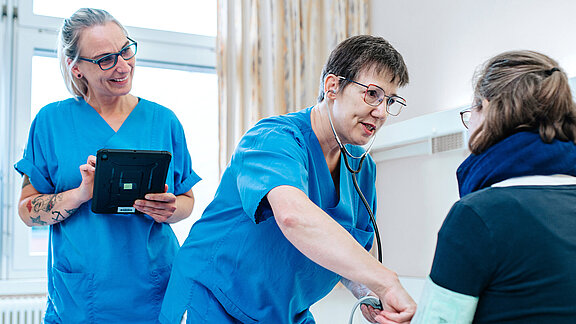 Zwei Pflegefachkräfte stehen am PatientInnenbett, die Patientin sitzt im Bett, eine Pflegefachkraft misst Blutdruck.