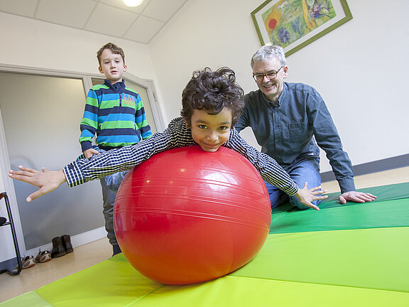 Physiotherapeut Sven Olde mit zwei Kindern in der Therapiesituation auf einer Gymnastikmatte, ein Kind liegt bäuchlings auf einem roten Gymnastikball, das zweite Kind hält die Füße des ersten Kindes.