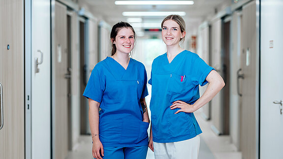 Zwei lächelnde Pflegefachkräfte, im Hintergrund der Krankenhausflur