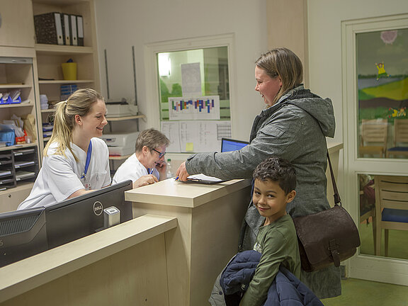 Eine Mutter mit Kind steht am Empfangstresen der Kinderstation.
