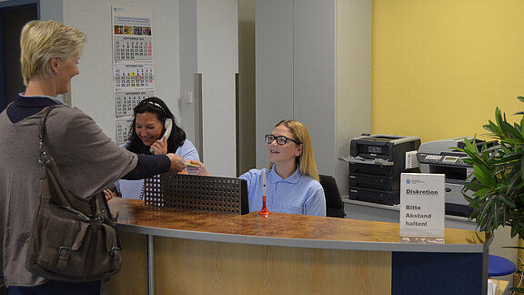 Eine Patientin am Anmeldetresen der HNO-Praxis, eine Mitarbeitende begrüßt die Patientin, die andere Mitarbeitende telefoniert.