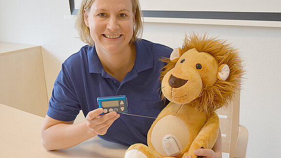 Oberärztin Nadine Scheffler hält ein Blutzuckermessgerät in der Hand, neben ihr am Tisch sitzt ein Kuscheltier (Löwe).