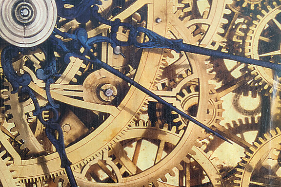 Zahnräder einer Uhr greifen ineinander, darüber sind blaue Zeiger abgebildet.