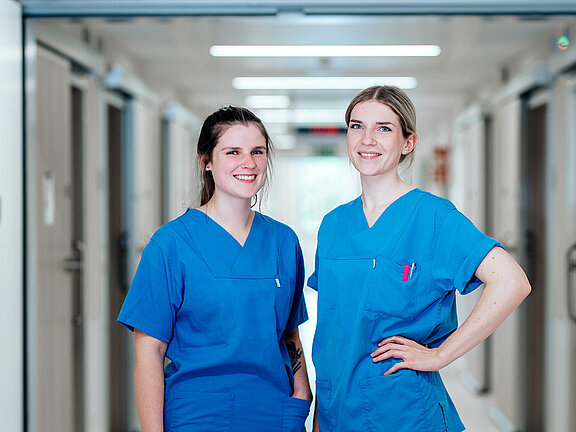 Zwei lächelnde Pflegefachkräfte, im Hintergrund der Krankenhausflur