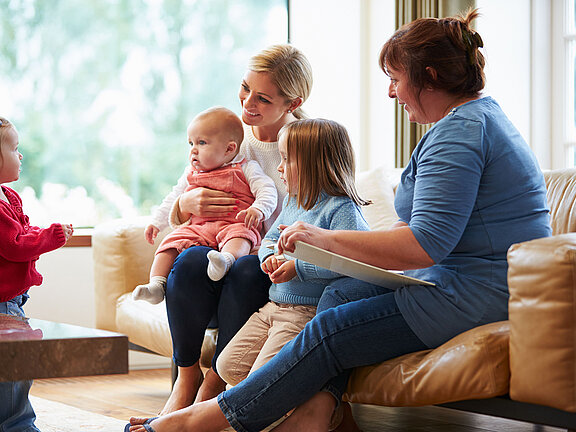 Zwei Frauen sitzen mit drei Kindern unterschiedlichen Alters in einem hellen Wohnzimmer