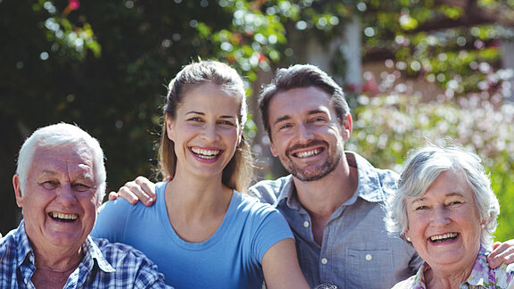 Eine fröhliche Familie bestehend aus Großeltern, Eltern und Kindern sitzen zusammen im Grünen und lächeln in die Kamera.