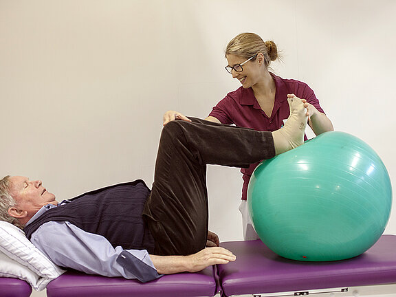 Eine Physiotherapeutin mobilisiert einen Patienten mit Hilfe eines Gymnastikballs